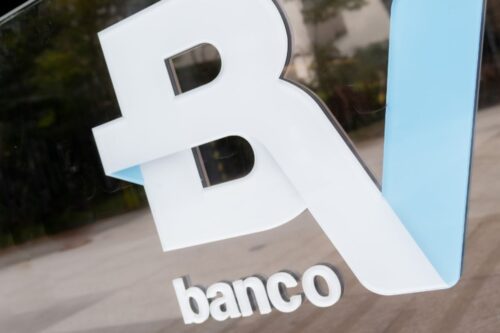 Logotipo em relevo do Banco BV fixado em uma placa de vidro transparente.