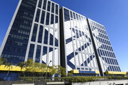Fachada de uma agência do Banco do Brasil com o logotipo amarelo e azul em destaque sobre uma estrutura metálica. O ambiente sugere uma unidade de atendimento em centro urbano, com vidraças e sinalização institucional.
