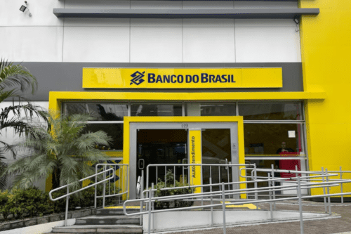 Fachada de uma agência do Banco do Brasil com as cores amarelo e cinza. A entrada possui rampas de acessibilidade com corrimãos metálicos e uma porta de vidro com a inscrição "Autoatendimento". À esquerda, há uma palmeira de pequeno porte.
