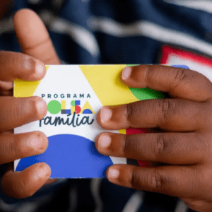 Close das mãos de uma criança de pele negra segurando o cartão do Programa Bolsa Família. O cartão é colorido, com formas geométricas em azul, amarelo e vermelho. No centro, lê-se "PROGRAMA BOLSA Família" com uma tipografia estilizada. A criança veste uma roupa escura com listras brancas e detalhes em vermelho.