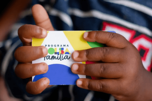 Close das mãos de uma criança de pele negra segurando o cartão do Programa Bolsa Família. O cartão é colorido, com formas geométricas em azul, amarelo e vermelho. No centro, lê-se "PROGRAMA BOLSA Família" com uma tipografia estilizada. A criança veste uma roupa escura com listras brancas e detalhes em vermelho.