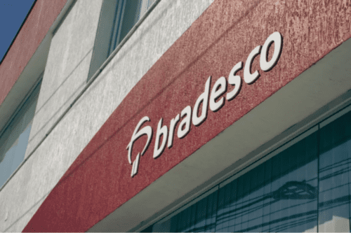 Fachada de uma agência bancária com destaque para o letreiro do Bradesco em letras brancas sobre uma parede de textura vermelha. A imagem está em ângulo diagonal, mostrando parte da estrutura do prédio e janelas de vidro.