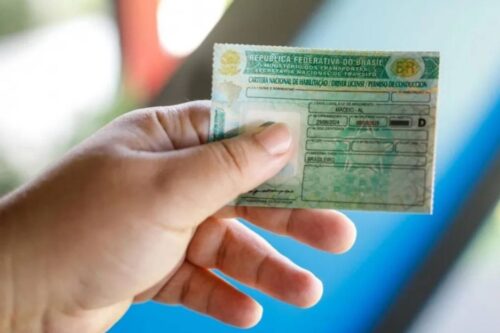 Uma mão segura em primeiro plano uma Carteira Nacional de Habilitação (CNH) brasileira física.