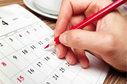 Close-up das mãos de uma pessoa segurando um lápis vermelho e marcando uma data em um calendário de papel sobre uma mesa de madeira.