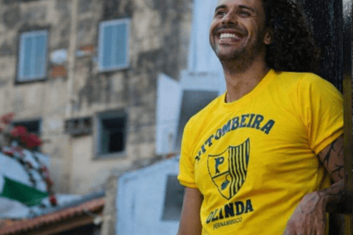 Homem jovem de cabelos cacheados e sorriso aberto, vestindo a tradicional camiseta amarela do bloco carnavalesco 'Pitombeira dos Quatro Cantos'. A estampa traz o escudo do bloco em azul e a inscrição 'Olinda - Pernambuco'. Ao fundo, vê-se o cenário histórico de Olinda com sobrados antigos desfocados.