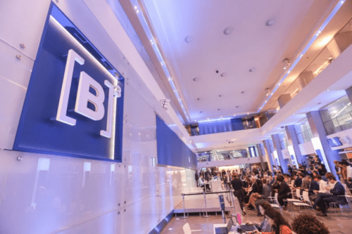 Visão ampla do saguão interno da B3, com o logotipo '[B]³' em destaque em um painel azul iluminado à esquerda. O ambiente é moderno, com pé-direito alto e iluminação clara, mostrando uma plateia sentada e profissionais trabalhando em um evento ou pregão.