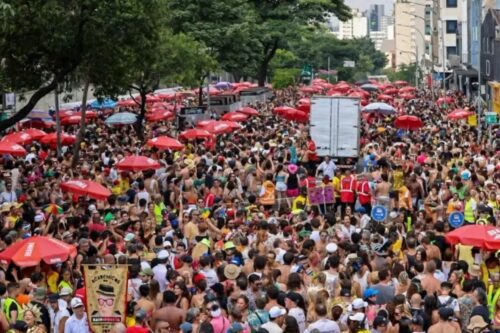 Vista aérea de uma multidão densa ocupando uma avenida arborizada durante o Carnaval de rua, com muitos guarda-sóis vermelhos de patrocinador espalhados pelo público.