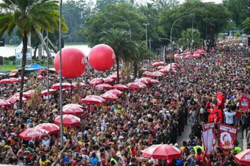 Fotografia em ângulo elevado mostrando uma multidão no Carnaval de rua em São Paulo, próxima ao Parque Ibirapuera, com balões vermelhos gigantes e guarda-sóis de marca.