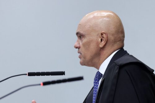 Fotografia de perfil do ministro Alexandre de Moraes. Ele está falando em um microfone, veste toga preta de magistrado sobre camisa branca e gravata azul estampada. O fundo é cinza claro e neutro.