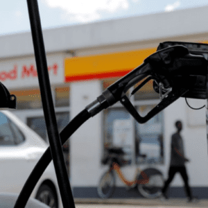 Close-up de um bico de abastecimento de combustível inserido no tanque de um veículo. Ao fundo, desfocado, vê-se a conveniência de um posto Shell.