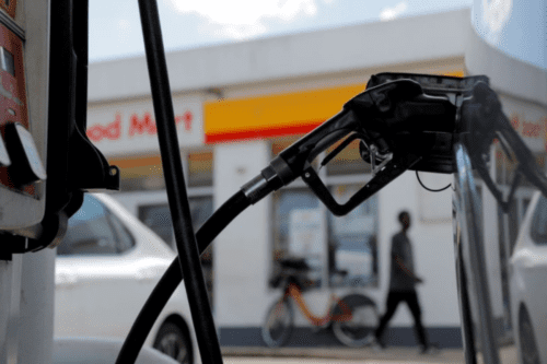 Close-up de um bico de abastecimento de combustível inserido no tanque de um veículo. Ao fundo, desfocado, vê-se a conveniência de um posto Shell.