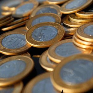 Close-up de diversas moedas de um real brasileiro (R$) empilhadas e espalhadas, destacando o brilho dourado e prateado do metal.