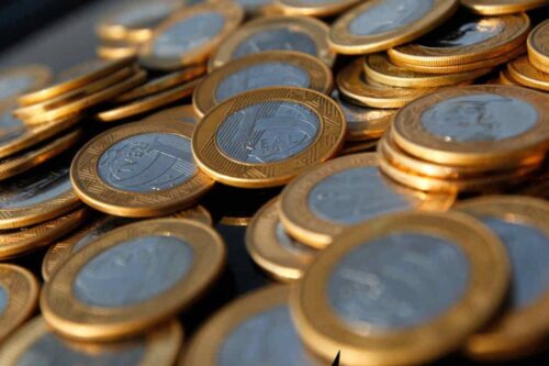 Close-up de diversas moedas de um real brasileiro (R$) empilhadas e espalhadas, destacando o brilho dourado e prateado do metal.