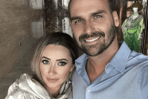 Uma selfie em plano fechado de Heloísa Bolsonaro e Eduardo Bolsonaro. Heloísa, à esquerda, tem cabelos loiros, olhos claros e usa uma maquiagem leve com batom rosado, vestindo um lenço ou echarpe de seda em tons claros. Eduardo, à direita, sorri para a câmera, possui barba curta e bem aparada e veste uma camisa social azul clara. Ao fundo, o ambiente parece ser o interior de um hotel ou loja, com uma parede de mármore e um manequim ao fundo vestindo um traje verde e chapéu.