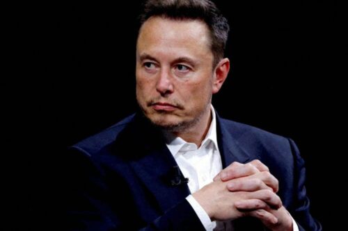 Elon Musk em um plano médio, vestindo um paletó azul escuro e camisa branca, com as mãos entrelaçadas e expressão séria contra um fundo preto.