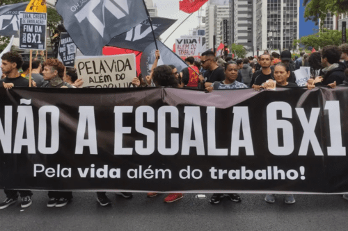 Fotografia de um protesto de rua em uma grande avenida urbana com prédios altos ao fundo. Manifestantes seguram uma grande faixa preta horizontal com letras brancas em destaque onde se lê: "NÃO À ESCALA 6X1 - Pela vida além do trabalho!". Ao fundo, outras pessoas carregam cartazes menores e bandeiras pretas e vermelhas. O clima é de mobilização social, com foco na pauta de direitos trabalhistas e redução da jornada de trabalho.