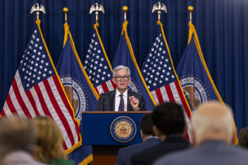 Jerome Powell, presidente do Federal Reserve, fala em um púlpito azul com o selo oficial do 'Board of Governors'. Ele usa terno escuro e óculos, gesticulando com a mão direita. Ao fundo, seis bandeiras dos Estados Unidos estão alinhadas sobre uma cortina azul escura.