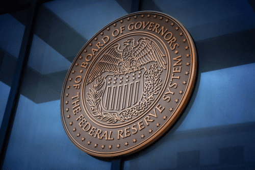 A imagem mostra em ângulo o selo oficial em relevo do Board of Governors of the Federal Reserve System (Banco Central dos Estados Unidos), fixado em uma superfície de vidro azulado.
