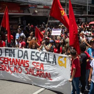 Manifestantes seguram uma grande faixa branca com letras pretas e vermelhas onde se lê: "Pelo fim da escala 6x1, 30h semanais sem redução salarial". Ao redor, há uma multidão com bandeiras vermelhas em uma rua comercial.