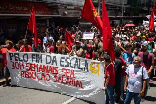 Manifestantes seguram uma grande faixa branca com letras pretas e vermelhas onde se lê: "Pelo fim da escala 6x1, 30h semanais sem redução salarial". Ao redor, há uma multidão com bandeiras vermelhas em uma rua comercial.