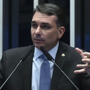 Fotografia em plano médio do senador Flávio Bolsonaro. Ele é um homem branco de cabelos curtos e escuros, veste terno preto, camisa azul clara e gravata azul com padrões geométricos. Ele está gesticulando com as duas mãos abertas enquanto fala em um púlpito com dois microfones à sua frente.