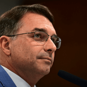 Retrato de perfil do senador Flávio Bolsonaro. Ele é um homem de pele clara, cabelos castanhos curtos e usa óculos de grau com armação fina. Está vestido com um paletó azul e camisa branca, posicionado diante de um microfone em um ambiente de iluminação sóbria, sugerindo um contexto de pronunciamento ou audiência.
