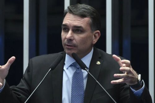 Fotografia em plano médio do senador Flávio Bolsonaro. Ele é um homem branco de cabelos curtos e escuros, veste terno preto, camisa azul clara e gravata azul com padrões geométricos. Ele está gesticulando com as duas mãos abertas enquanto fala em um púlpito com dois microfones à sua frente.