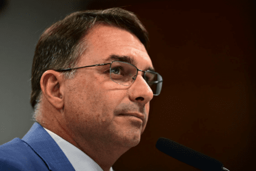 Retrato de perfil do senador Flávio Bolsonaro. Ele é um homem de pele clara, cabelos castanhos curtos e usa óculos de grau com armação fina. Está vestido com um paletó azul e camisa branca, posicionado diante de um microfone em um ambiente de iluminação sóbria, sugerindo um contexto de pronunciamento ou audiência.
