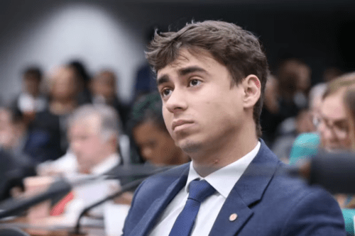 Retrato em close do deputado Nikolas Ferreira na Câmara dos Deputados. Ele tem cabelos castanhos curtos, veste um paletó azul-marinho e gravata azul texturizada sobre camisa branca. Ele olha para cima e para a esquerda com uma expressão séria e pensativa. O fundo está desfocado, mostrando outros parlamentares e microfones em um ambiente de comissão.