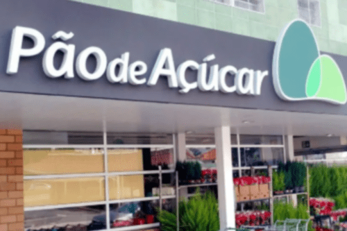 Fachada de uma unidade do supermercado Pão de Açúcar. O letreiro exibe o nome da marca em letras brancas sobre um painel cinza escuro, acompanhado pelo logotipo de dois semicírculos verdes à direita. Abaixo, é possível ver a entrada da loja com plantas ornamentais.
