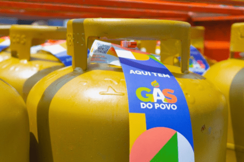 Close-up de vários botijões de gás de cozinha na cor dourada, enfileirados. O botijão em primeiro plano possui um rótulo adesivo colorido (azul, amarelo, verde e rosa) com a frase "AQUI TEM GÁS DO POVO" e um pequeno QR Code no topo. Ao fundo, outros botijões similares aparecem levemente desfocados sob uma estrutura metálica laranja.