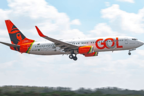 Vista lateral de um avião da companhia aérea GOL Linhas Aéreas em voo, prestes a pousar. A aeronave é branca com o logotipo "GOL" em laranja na fuselagem. A cauda é personalizada com uma ilustração em preto e laranja que homenageia Santos Dumont, celebrando os "150 anos" do aviador. O céu ao fundo está claro com poucas nuvens.