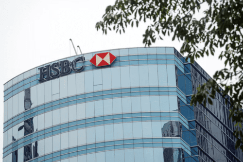 Detalhe da parte superior de um moderno edifício comercial com fachada curva de vidro espelhado azulado. No topo, destaca-se o logotipo do banco HSBC: a sigla em letras escuras ao lado de um símbolo de hexágono estilizado em vermelho e branco. No canto superior direito, galhos de árvores com folhas verdes emolduram a cena contra um céu claro.
