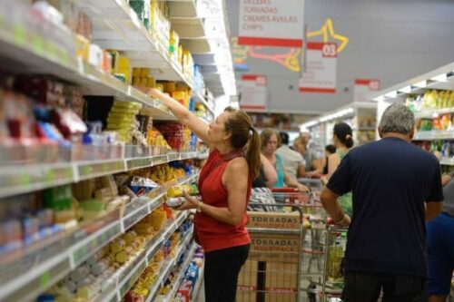 Uma mulher de blusa vermelha observa produtos em uma prateleira de supermercado lotada.