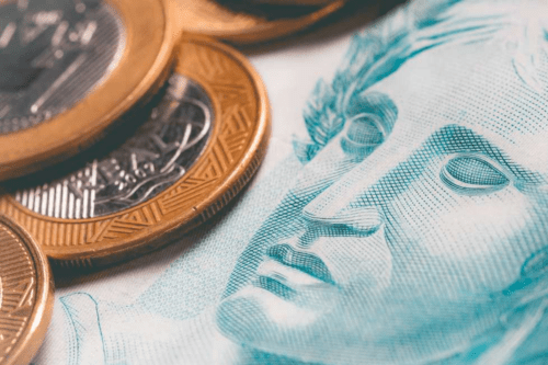 Uma composição em close-up de dinheiro brasileiro. No lado esquerdo, várias moedas de um Real estão empilhadas e espalhadas; elas possuem o anel externo dourado e o centro prateado. No lado direito, em destaque e levemente fora de foco, aparece parte de uma cédula de Real em tons de azul, mostrando o rosto da Efígie da República. A iluminação é suave, criando um aspecto editorial sobre o tema de economia.