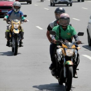 Trânsito urbano em uma avenida larga sob luz solar forte. Em destaque, motociclistas pilotando suas motos entre os carros; um deles veste uma camisa verde e leva um passageiro.