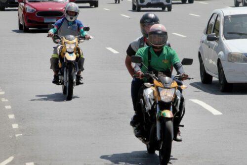 Trânsito urbano em uma avenida larga sob luz solar forte. Em destaque, motociclistas pilotando suas motos entre os carros; um deles veste uma camisa verde e leva um passageiro.