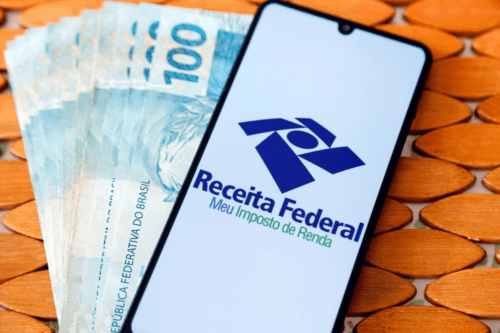 Um smartphone em primeiro plano exibe o logotipo da Receita Federal com a frase 'Meu Imposto de Renda'. Ao fundo, notas de 100 reais dispostas em leque sobre uma superfície de madeira texturizada