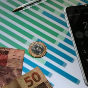 Composição sobre finanças brasileiras mostrando notas de 100 e 50 reais, uma moeda de 1 real sobre um gráfico de barras coloridas, e um smartphone ao lado exibindo o aplicativo de calculadora com o número 2.021 na tela.