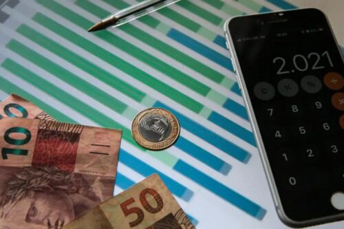 Composição sobre finanças brasileiras mostrando notas de 100 e 50 reais, uma moeda de 1 real sobre um gráfico de barras coloridas, e um smartphone ao lado exibindo o aplicativo de calculadora com o número 2.021 na tela.