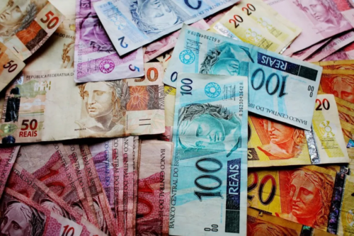 Diversas cédulas de Real (BRL) de diferentes valores, como notas de 100, 50, 20 e 10 reais, espalhadas de forma sobreposta, representando o cenário econômico e investimentos no Brasil.