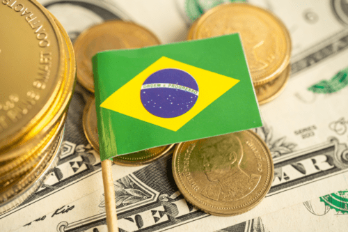 Notas e moedas brasileiras ao redor de uma pequena bandeira do Brasil sobre uma nota de dólar.