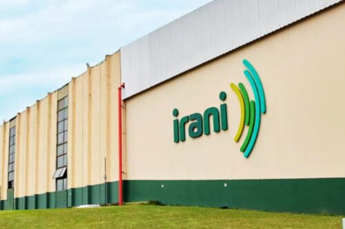 Fachada lateral de uma unidade industrial da empresa Irani. A parede bege exibe o logotipo da marca em verde e amarelo, com um gramado em primeiro plano e o topo do edifício em zinco sob um céu claro.