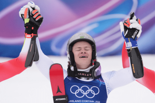Um atleta de esportes de inverno comemora com entusiasmo nos Jogos de Milano Cortina 2026. Ele veste um traje de competição branco e azul com os anéis olímpicos no peito e um capacete cinza com a bandeira do Brasil. O atleta está com os olhos fechados e a boca aberta em um grito de vitória, enquanto seus braços são levantados por outras pessoas (visíveis apenas os braços e luvas). Ele segura um esqui vermelho da marca Atomic. O fundo é composto por neve e tons de roxo e azul.