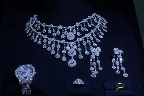 Conjunto de joias de luxo prateadas cravejadas de diamantes ou pedras brilhantes sobre um mostruário de veludo preto. O conjunto inclui um colar de duas voltas com diversos pingentes em formato de gota, um par de brincos longos, um anel e um relógio de pulso com pulseira e mostrador também cobertos por pedras. No canto inferior direito, há uma pequena chave dourada.