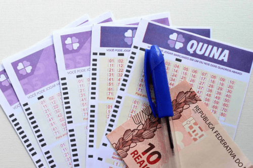 Imagem de várias folhas de jogo da Quina com um bilhete de R$10 ao centro e uma caneta azul, representando apostas na loteria.