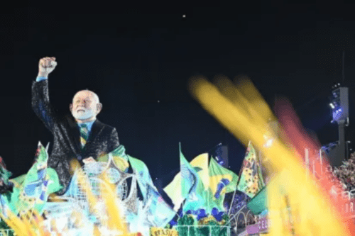 Um Boneco do presidente Lula, vestindo um paletó escuro brilhante e gravata colorida, está no alto de um carro alegórico durante o desfile de Carnaval. Ele tem o braço direito erguido com o punho cerrado. Ao redor, veem-se bandeiras do Brasil e luzes vibrantes de cor amarela e verde em movimento, típicas da Sapucaí.