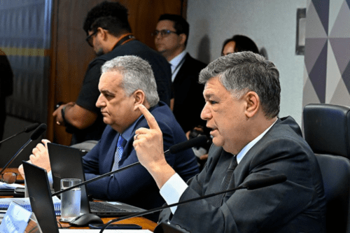 Dois homens em trajes formais sentados em uma mesa de conferência com microfones e laptops; o homem em primeiro plano gesticula com o dedo indicador levantado durante uma fala.