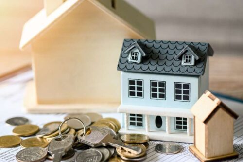 otografia em close-up de miniaturas de casas de diferentes estilos sobre uma mesa, cercadas por moedas e chaves de metal, simbolizando o mercado imobiliário e financiamento habitacional.