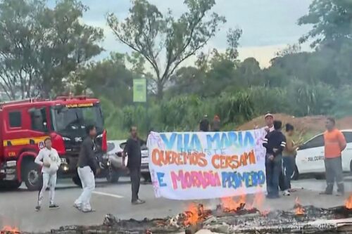 Manifestantes realizam um protesto em uma rodovia, segurando uma grande faixa artesanal onde se lê: "VILA MARIA RESISTE. QUEREMOS CERSAM E MORADIA PRIMEIRO".